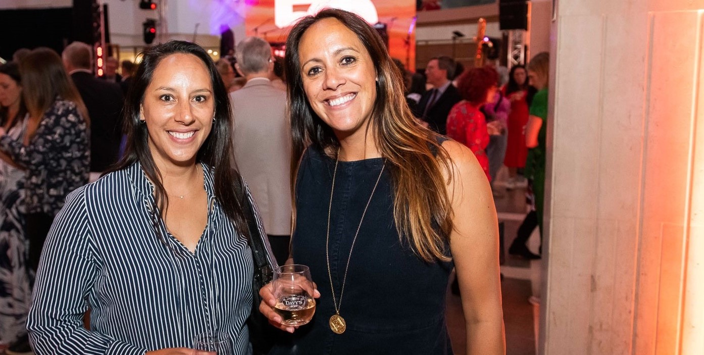 Two women smile at the camera against the backdrop of a party at the British Library 